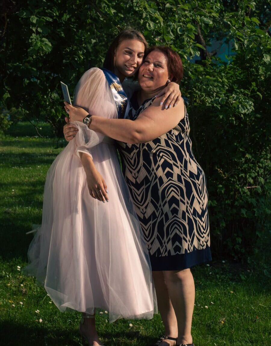 A smiling mother embraces her daughter, the bride, in a lush outdoor setting, radiating happiness and love.